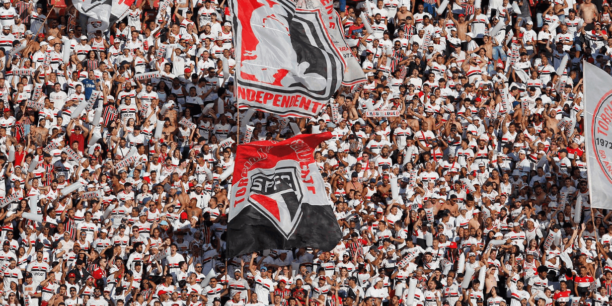 Torcida do São Paulo Futebol Clube vestida de branco e vermelho, agitando bandeiras e cantando nas arquibancadas do Estádio do Morumbi.